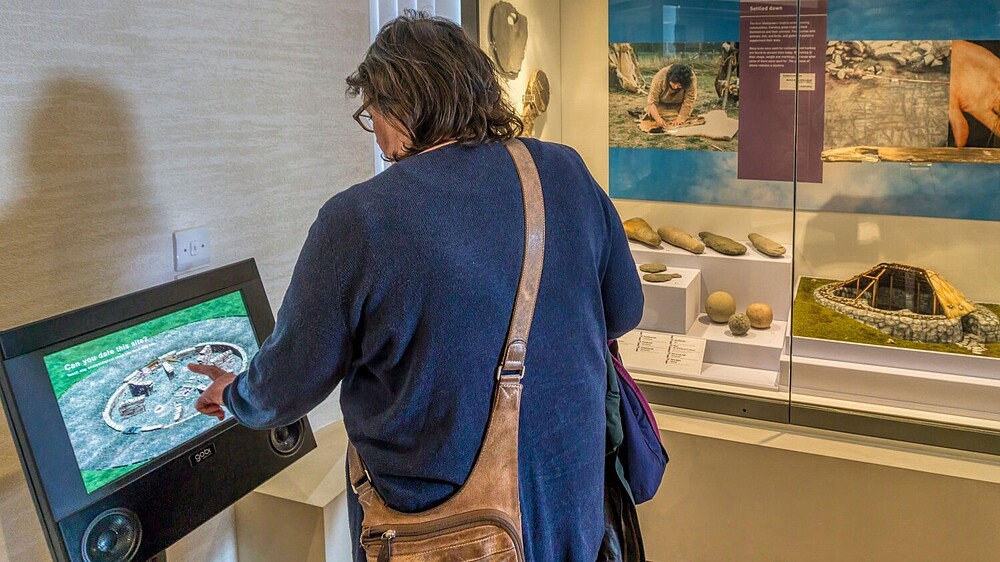 © UrbanImages / Alamy Stock Foto A museum visitor stands in front of a showcase with ancient objects and uses a touchscreen standing next to it. © UrbanImages / Alamy Stock Foto