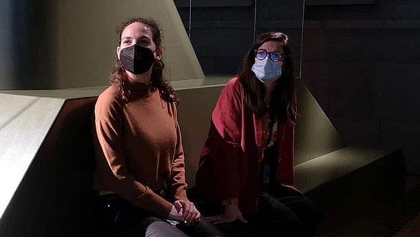  Two people sit in a dark exhibition space. They wear masks and look towards the left of the picture.