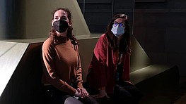 Two people sit in a dark exhibition space. They wear masks and look towards the left of the picture.