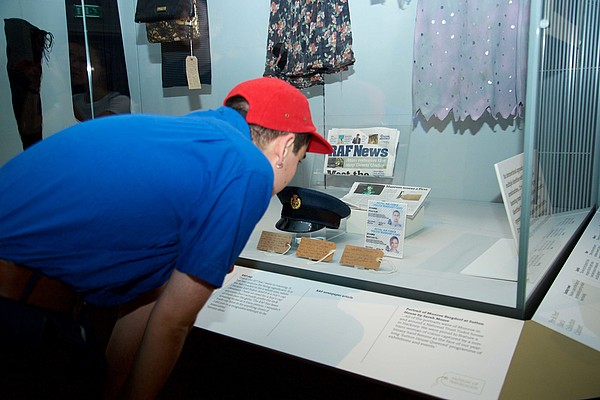 Person standing with their back to the photographer in blue shirt and red cap leans over to look at objects in a display case.