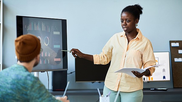 A person stands next to a screen and gives a presentation to another person, who sits at a desk.