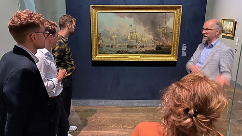 A group of people are photographed from behind looking at a painting of a ship in a golden frame. One person is turned towards the group and talks to them.  