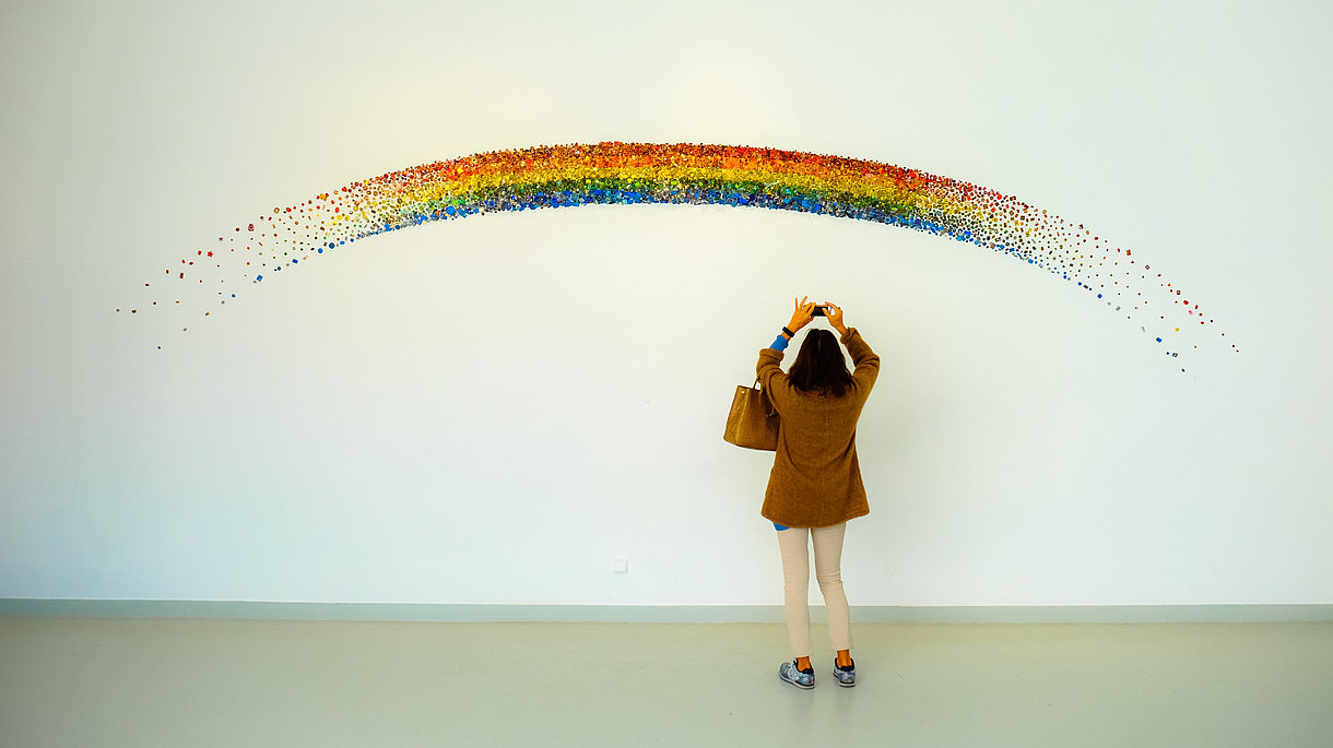 © Image: Francesco Ungaro A person reaches up to take a photo with their phone of a rainbow that is painted on a wall. 