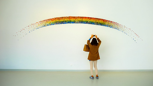  A person reaches up to take a photo with their phone of a rainbow that is painted on a wall. 