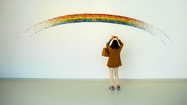 A person reaches up to take a photo with their phone of a rainbow that is painted on a wall. 