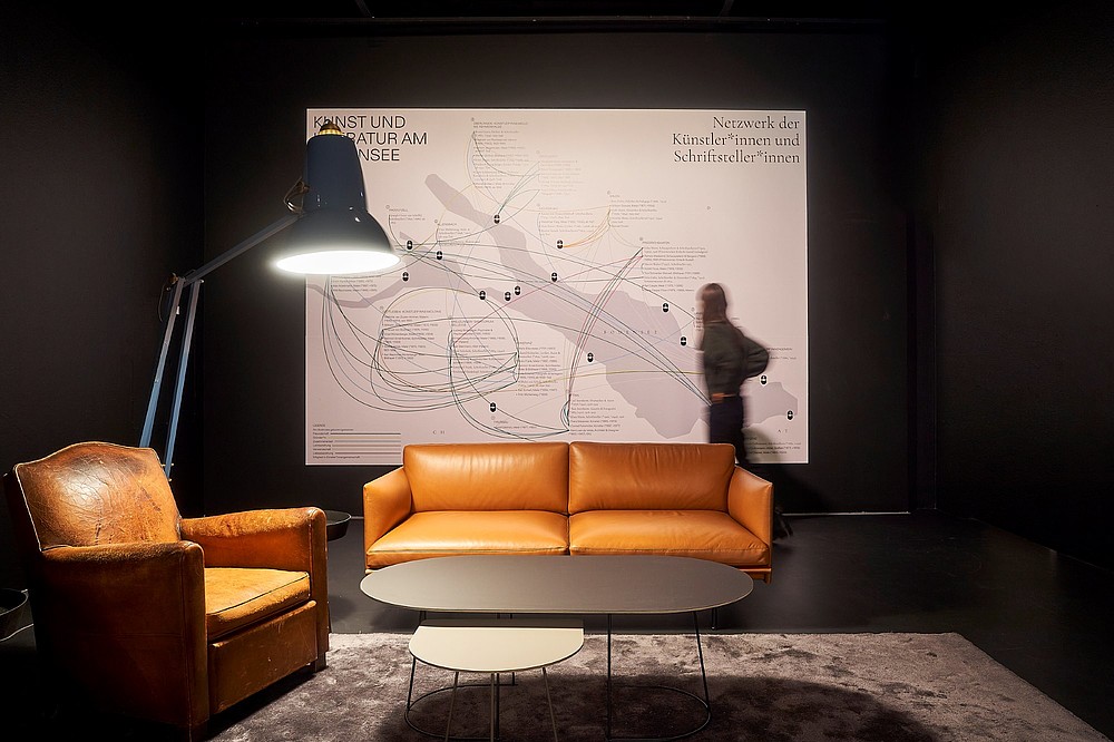 A person looks at a large exhibition poster. In the foreground is a sofa, armchair and a big lamp.  