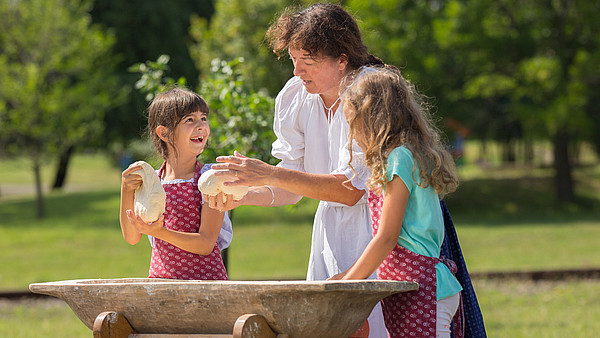 This photograph depicts a person showing to children how to handle dough. They stand outside behind a wooden trough.