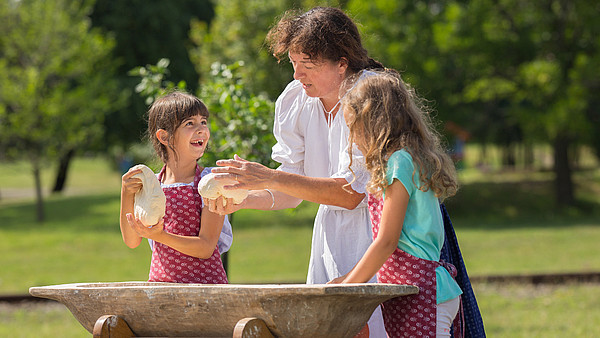 This photograph depicts a person showing to children how to handle dough. They stand outside behind a wooden trough.