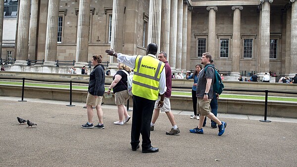 A security officer guides a group of people towards the left hand side of the photograph. In the background, people queing in front of a museum can be seen.