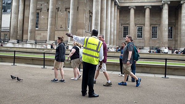 A security officer guides a group of people towards the left hand side of the photograph. In the background, people queing in front of a museum can be seen.