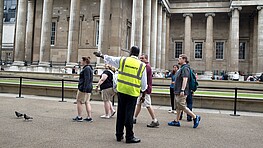 A security officer guides a group of people towards the left hand side of the photograph. In the background, people queing in front of a museum can be seen.