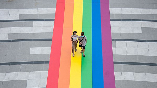 Two people, photogrtaphed from bird perspective, walk over a huge LGBTQ flat that has been attached to the floor.