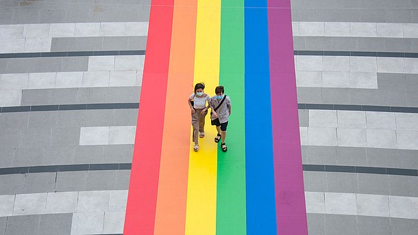  Two people, photogrtaphed from bird perspective, walk over a huge LGBTQ flat that has been attached to the floor.