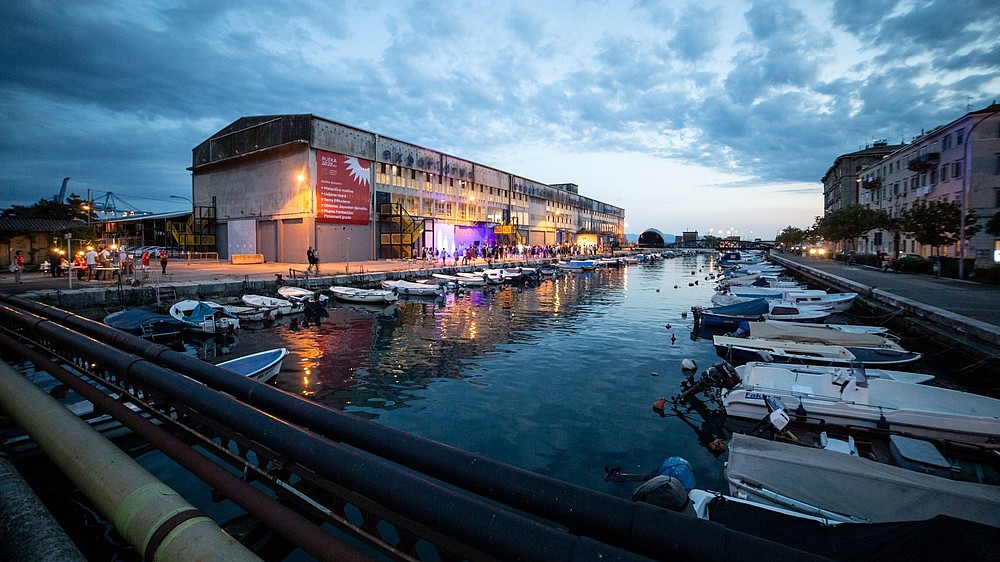 A harbour with little boats front of an evening sky. In the background is a large industrial looking building. Numerous people are standing in front of it.   
