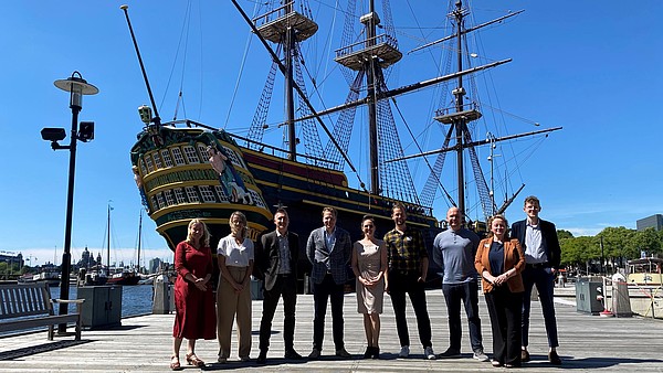 This photograph shows a group of people standing in front of a large ship on a pier. They are smiling into the camera. The sky is blue. 