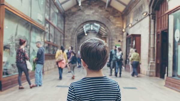 A child is photographed from behind. They are walking through an exhibition space where a few people are also looking at the displays.
