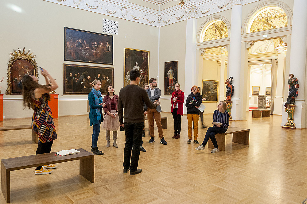 Photograph of a number of people standing in a circle in a museum. They look around towards the pictures and sculptures.  