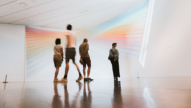  Photograph of people walking through an empty gallery space. They look at a rainbow coloured light installation
