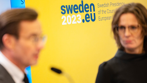  This photograph shows two people in the foreground who are out of focus. The background consists of a yellow wall and the logo of the Swedish Presidency of the Council of the European Union.