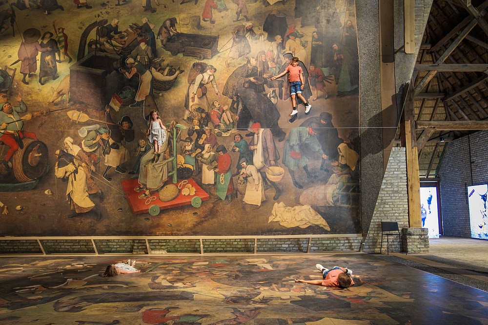Two kids lay down on a blown up version of a Bruegel painting. A huge tilted mirror shows them as part of the painting.  © Bokrijk