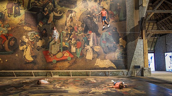 Two kids lay down on a blown up version of a Bruegel painting. A huge tilted mirror shows them as part of the painting.