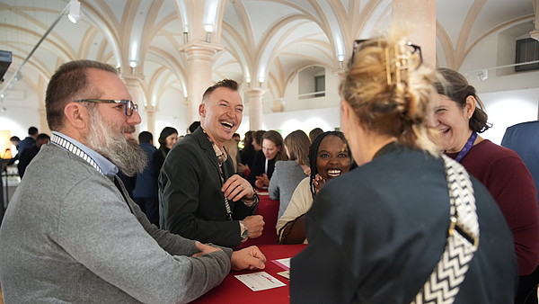 Four people stand around a table engaging in conversation and laughing. 