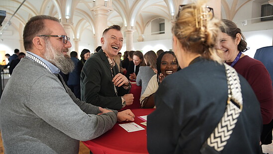 Four people stand around a table engaging in conversation and laughing. 