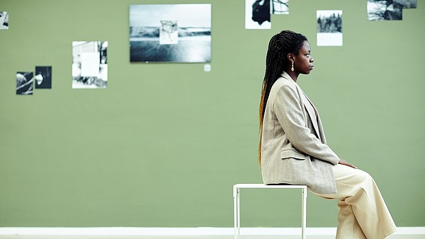 A person, photographed from the side, sits on a stool. The wall behind them is covered with black and white photographs.