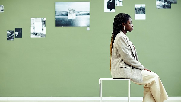 A person, photographed from the side, sits on a stool. The wall behind them is covered with black and white photographs.