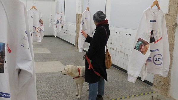 Person wearing a face mask is standing in a gallery together with her dog. She is looking at one of many exhibited sweatshirts that are hanging from the ceiling.
