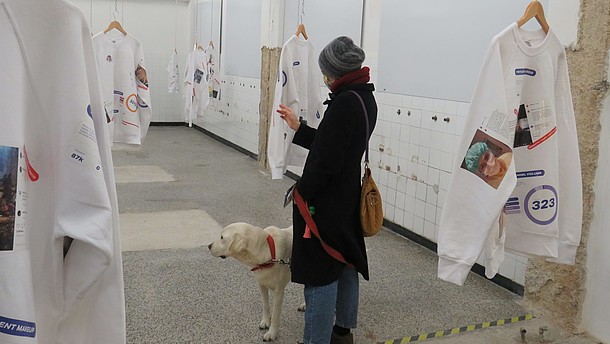  Person wearing a face mask is standing in a gallery together with her dog. She is looking at one of many exhibited sweatshirts that are hanging from the ceiling.