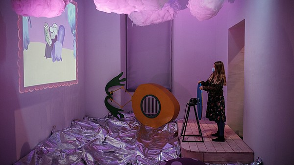 A person is standing in a pink lit exhibition display. They are holding a steering wheel and looking towards a projection.