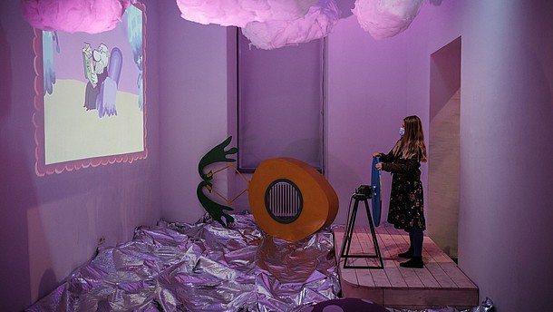  A person is standing in a pink lit exhibition display. They are holding a steering wheel and looking towards a projection.