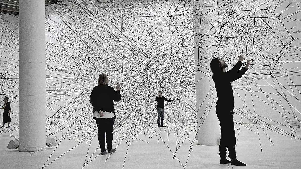  Black and white photograph of people in a white exhibition space. They interact with a spacious art piece made out of strings.