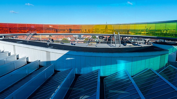  Rooftop overlooking a city with solar panels in the foreground of picture and big colourful see-through artwork in the background that people are walking through.