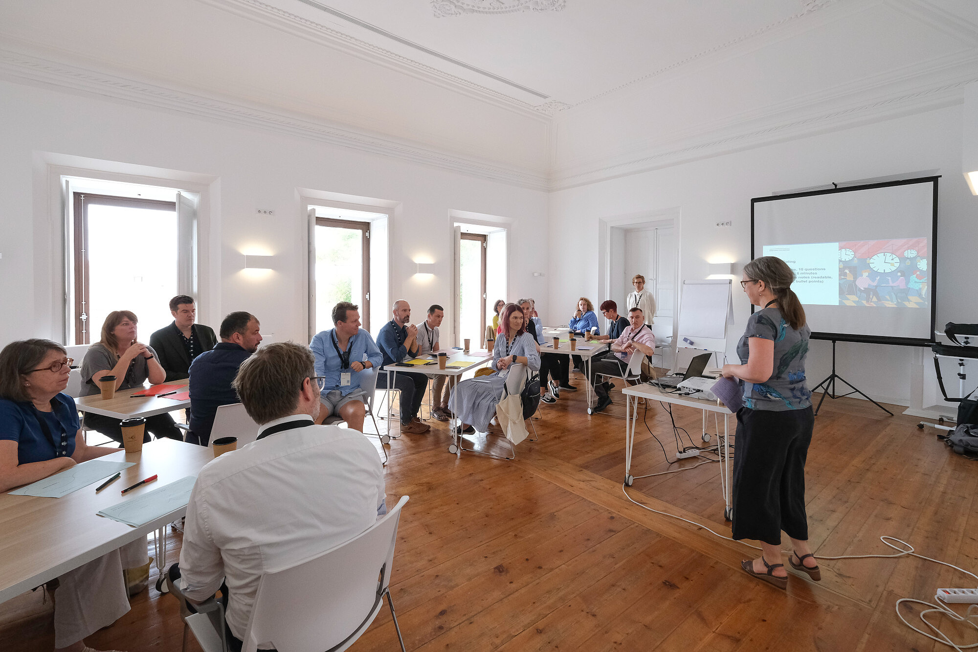 © Image: Jorge Gomes People are sitting on small tables in a conference room. One person is standing and talking. Presentation slides are projected to a screen at the front of the room. © Image: Jorge Gomes
