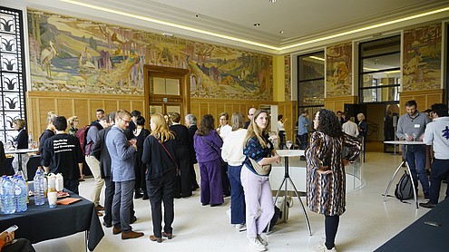 Photo of a group of people who are talking to each other in smaller groups while having refreshments. Only one person is looking into the camera. The room has decorative wall paintings of landscapes with birds and animals.  © Image: Peter Vand der Plaetsen