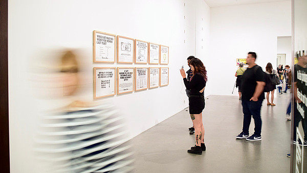 People are taking photos and look at paintings inside a gallery. A person in the foreground is leaving the room and is therefore blurry in the picture. 