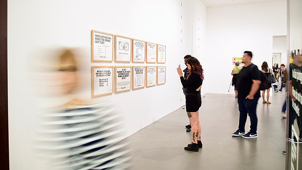  People are taking photos and look at paintings inside a gallery. A person in the foreground is leaving the room and is therefore blurry in the picture. 