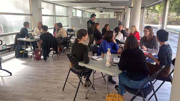Three groups of about 5-6 people each are seated around three tables each carrying out a workshop. The workshop leader is standing up discusses something with one of the groups. 