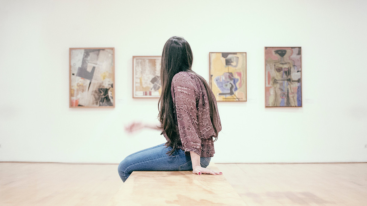 © Image: Gilber Franco A woman in profile is sitting down on a bench and looks at four paintings on a wall. 