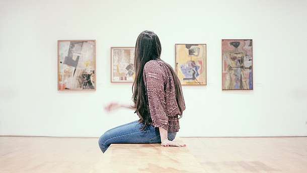  A woman in profile is sitting down on a bench and looks at four paintings on a wall. 