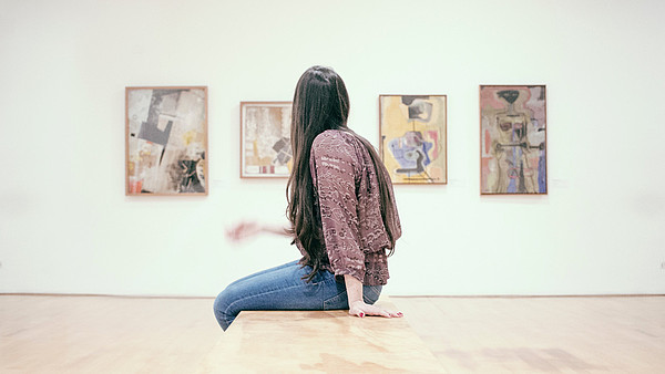 A woman in profile is sitting down on a bench and looks at four paintings on a wall. 
