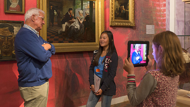  One girl is using a tablet to film another girl who is talking with an older man, presumably about the painting behind them.