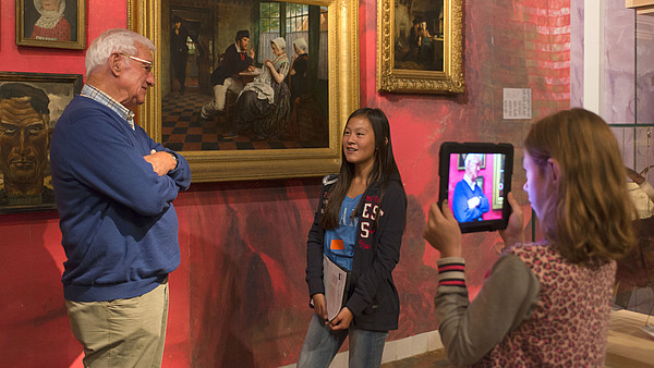 One girl is using a tablet to film another girl who is talking with an older man, presumably about the painting behind them.