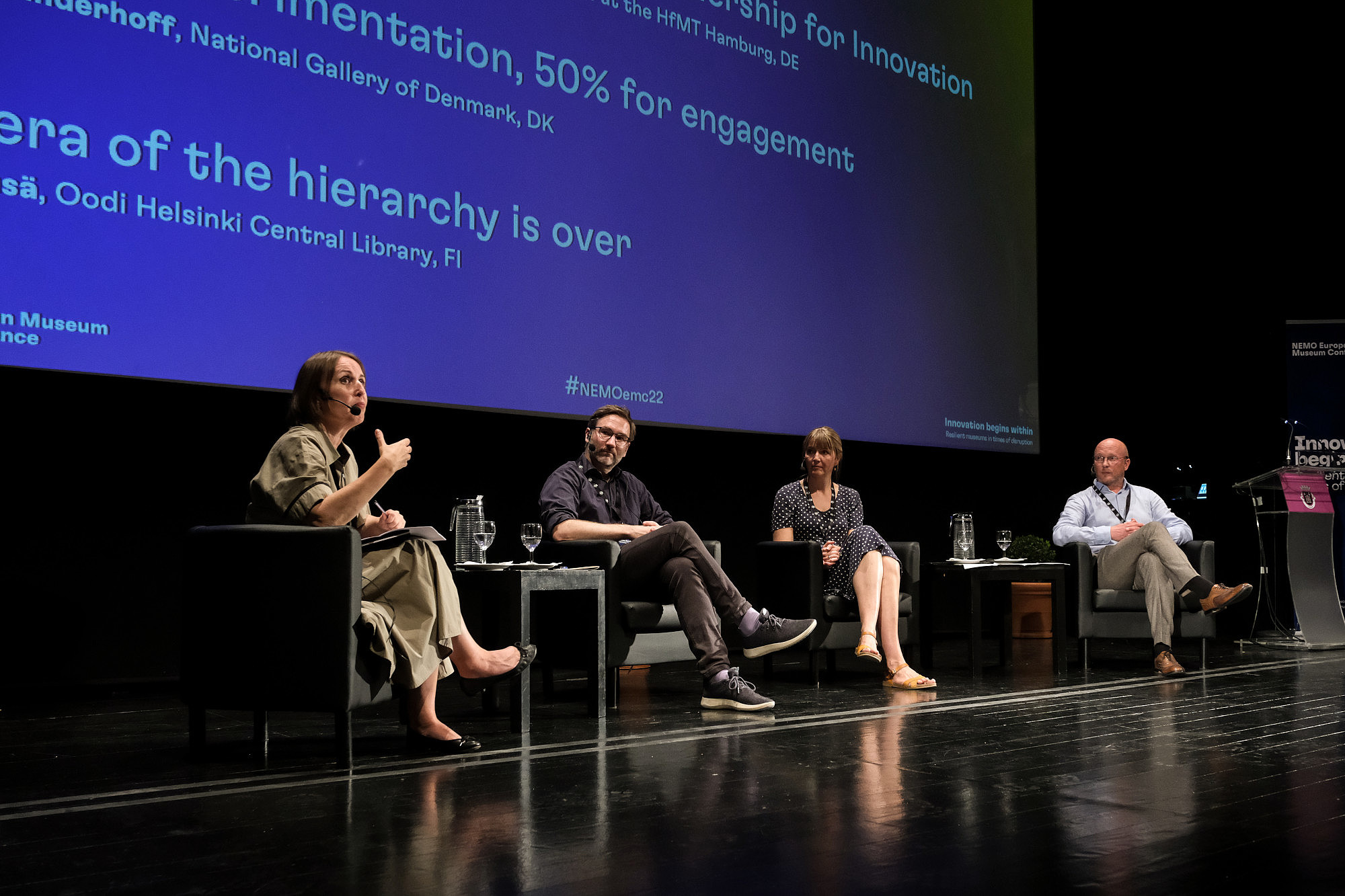 Four people are sitting on a stage. One person is talking, the others are looking at them.In the background, presentation slides are projected to a screen.   © Image: Jorge Gomes