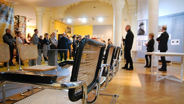 Two rows of chairs are facing each other in an exhibition space. In the background three people are giving a presentation to a group.