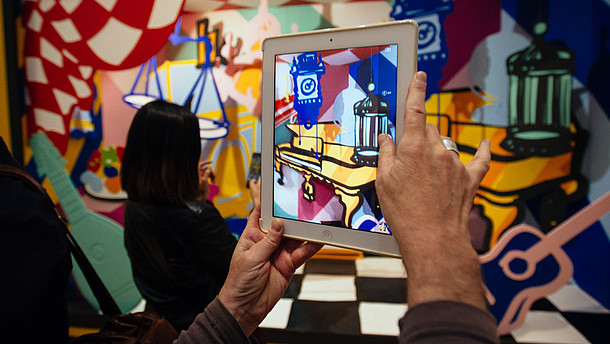  In this photograph a person takes a photo of a colourful and abstract installation using a tablet. The photo shows only their hands.