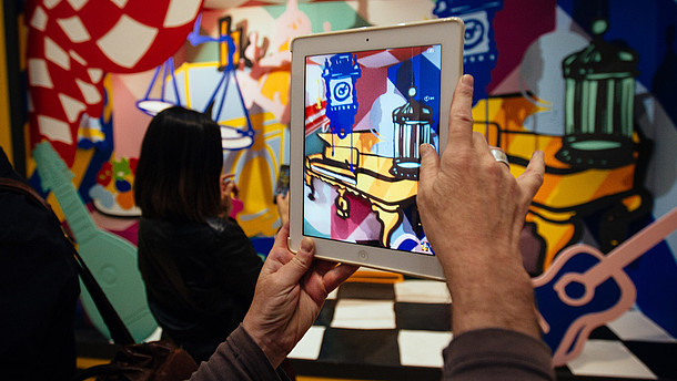  In this photograph a person takes a photo of a colourful and abstract installation using a tablet. The photo shows only their hands.