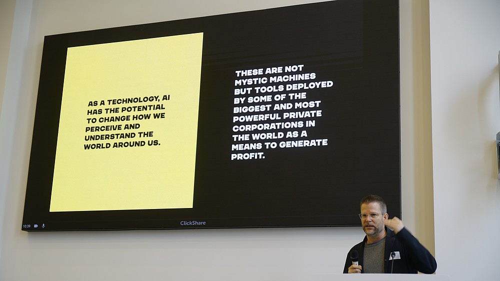A persons is seen speaking next to a projected image.   © Image: Peter Vand der Plaetsen